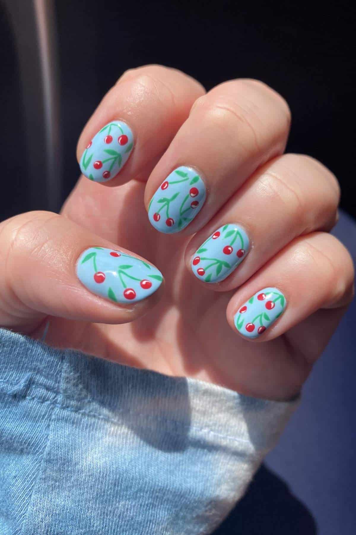 A hand with light blue nail polish featuring a cherry and green branch design on each fingernail.