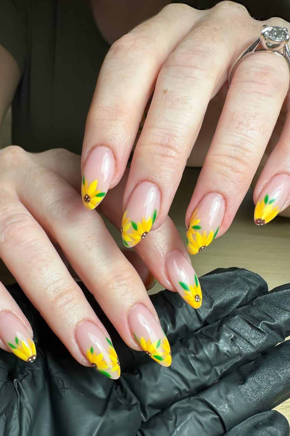 Hands with almond-shaped nails featuring yellow sunflower designs on the tips, over a neutral base. One hand wears a silver ring, the other rests on a black glove.
