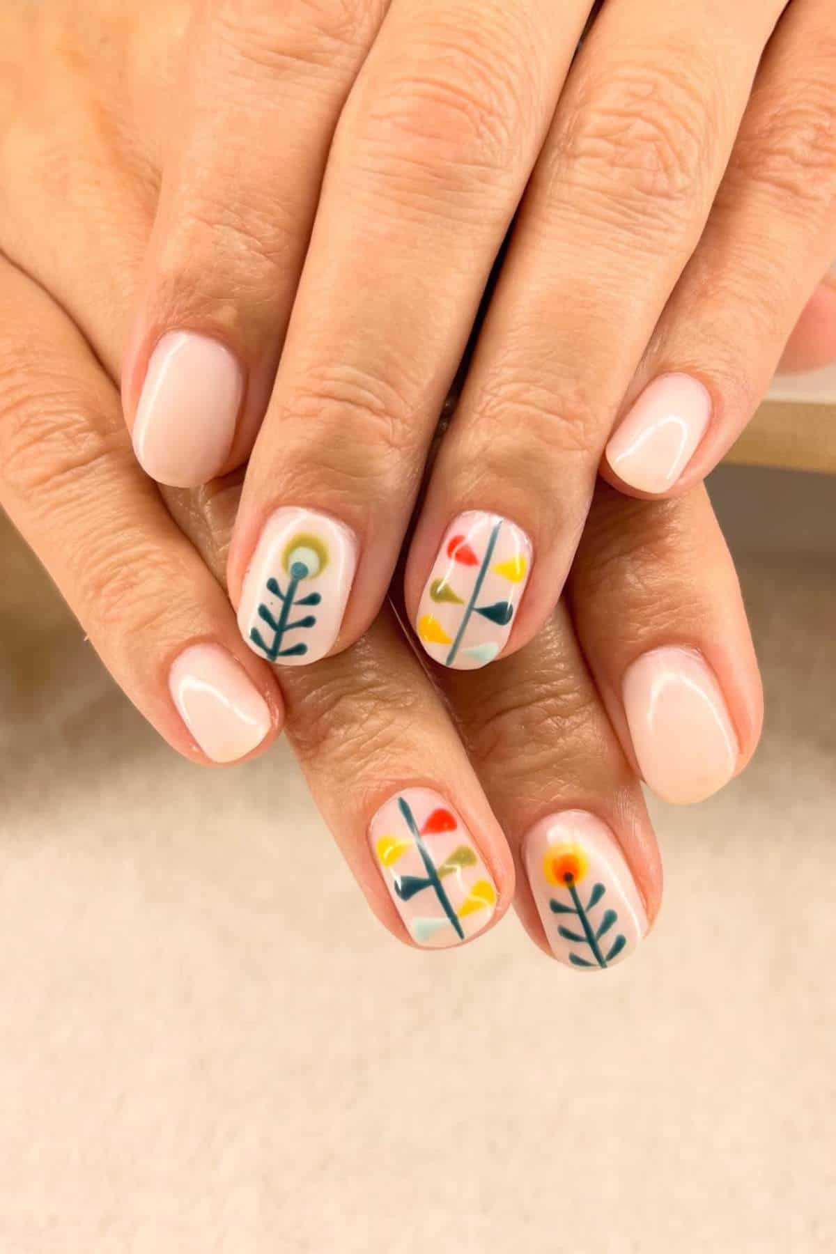 Hands with short, oval nails painted in a light nude polish; two nails on each hand feature minimalist, colorful floral designs with stems and leaves.