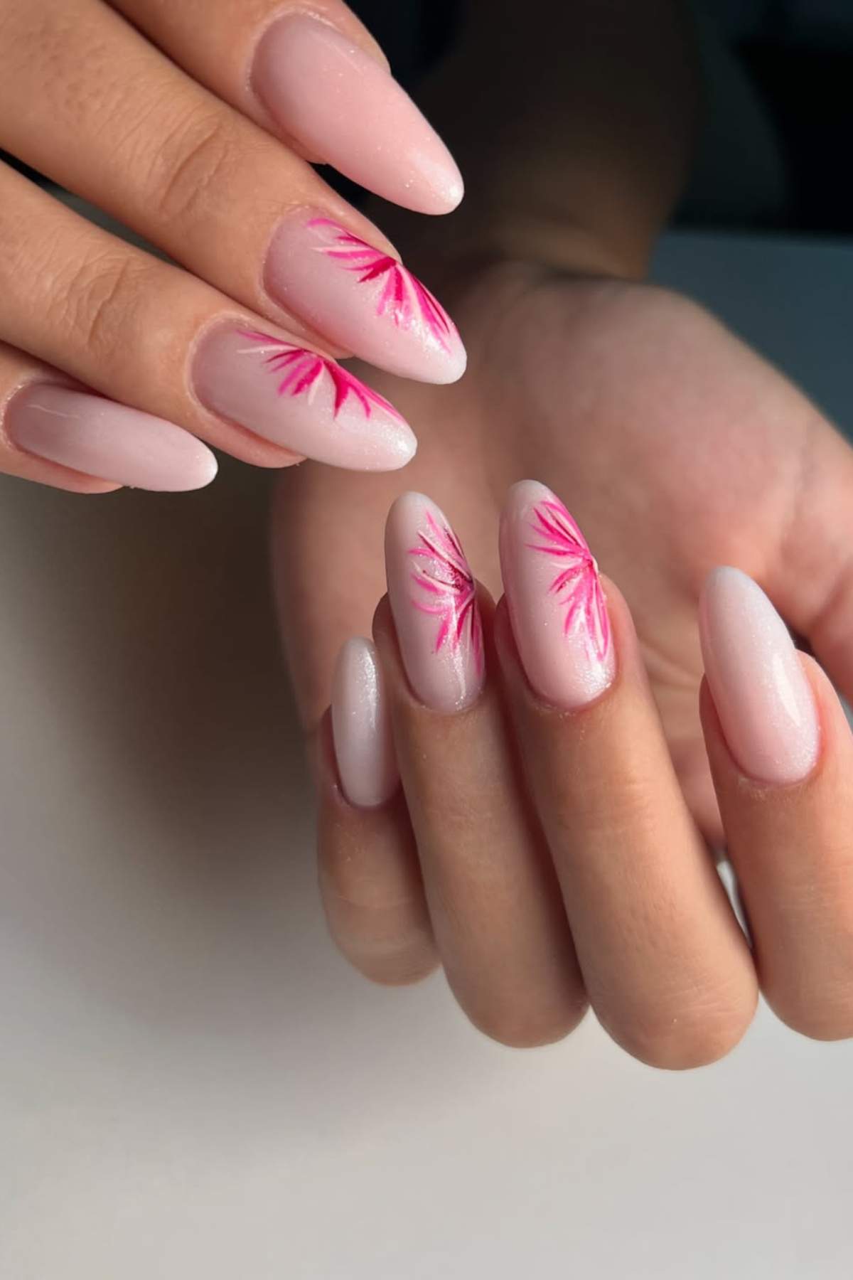 A hand with almond-shaped nails painted light pink, featuring pink leaf-like designs on two accent nails.