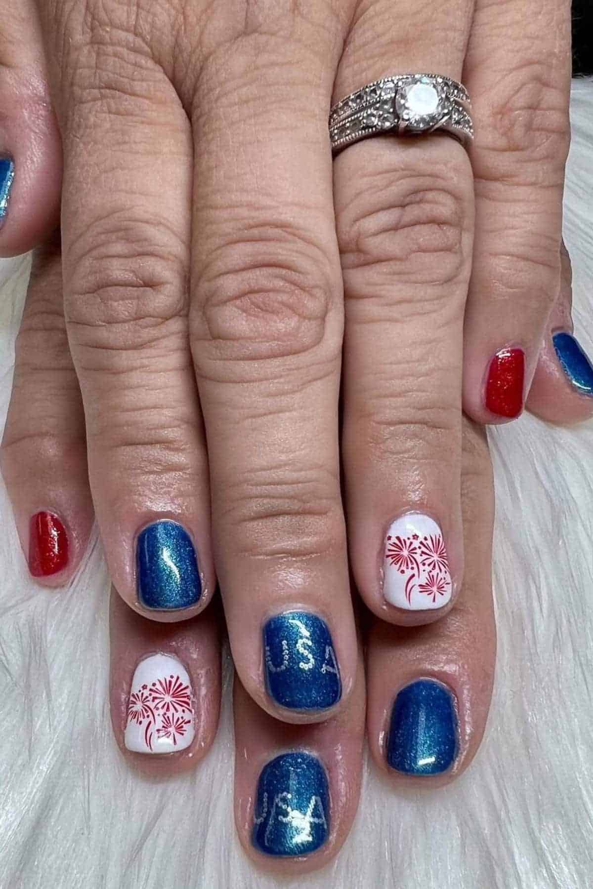 Hands with red, white, and blue nails featuring fireworks and "USA" designs. A silver ring with a diamond is visible on one finger.