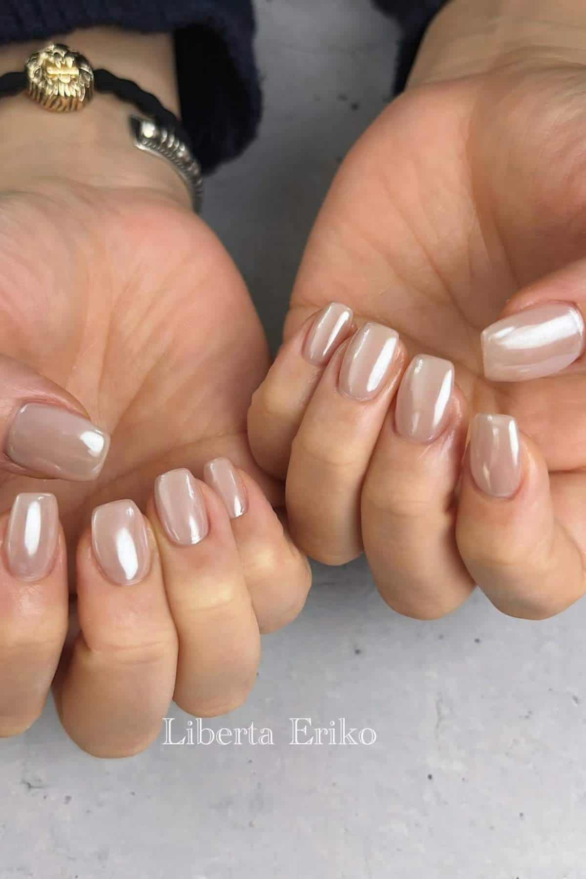 Close-up of two hands with short, square-shaped nails painted in a glossy nude polish. A bracelet is visible on one wrist. The background is a light gray surface.