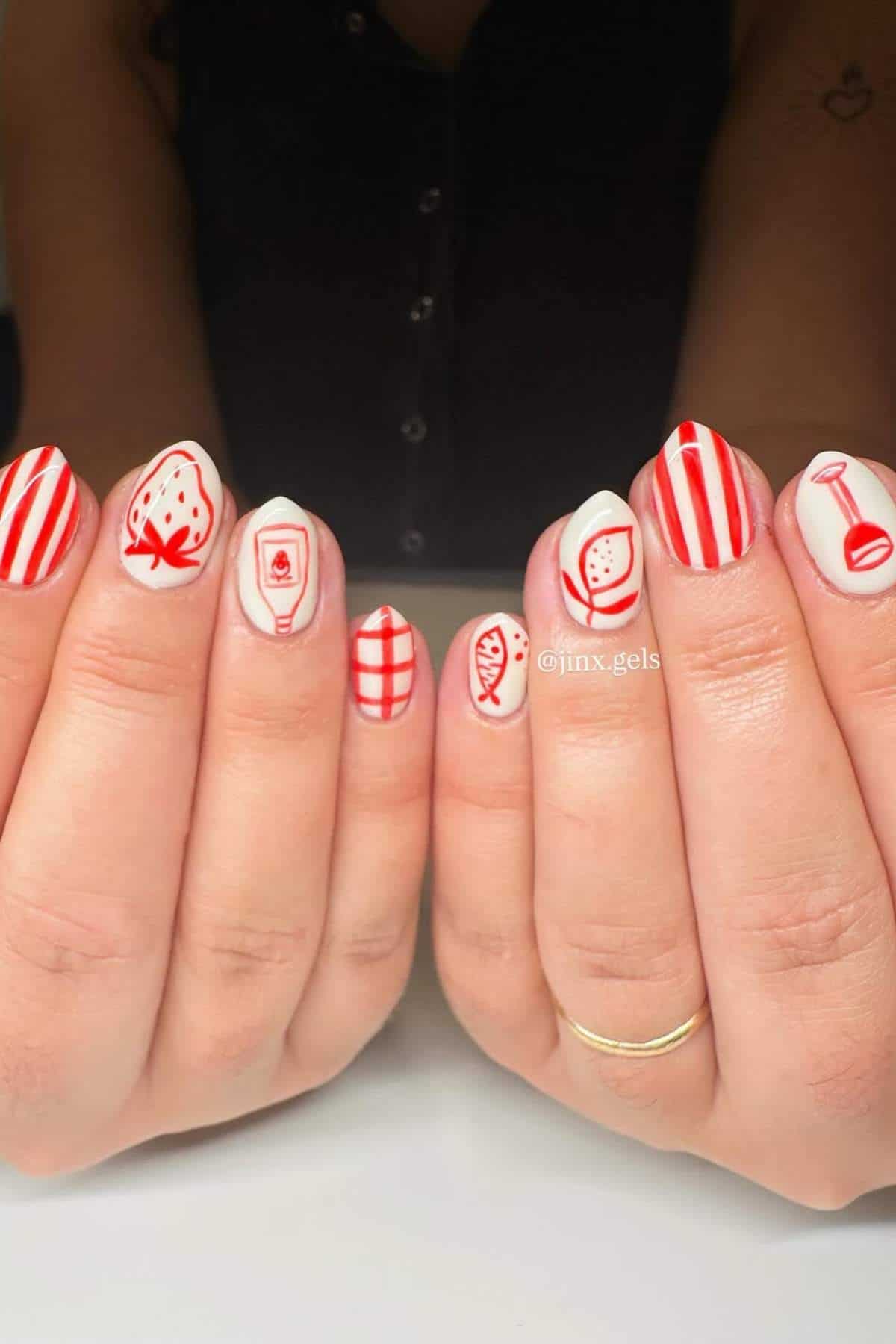 Close-up of hands with white nail polish and red designs, including stripes, a strawberry, a drink, a checkered pattern, a fish, and a cup.