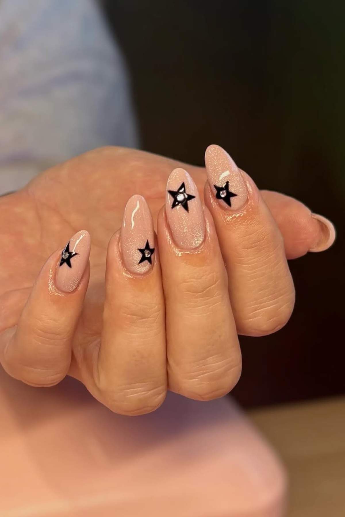 A hand with almond-shaped nude nails, each featuring a black star design as nail art.