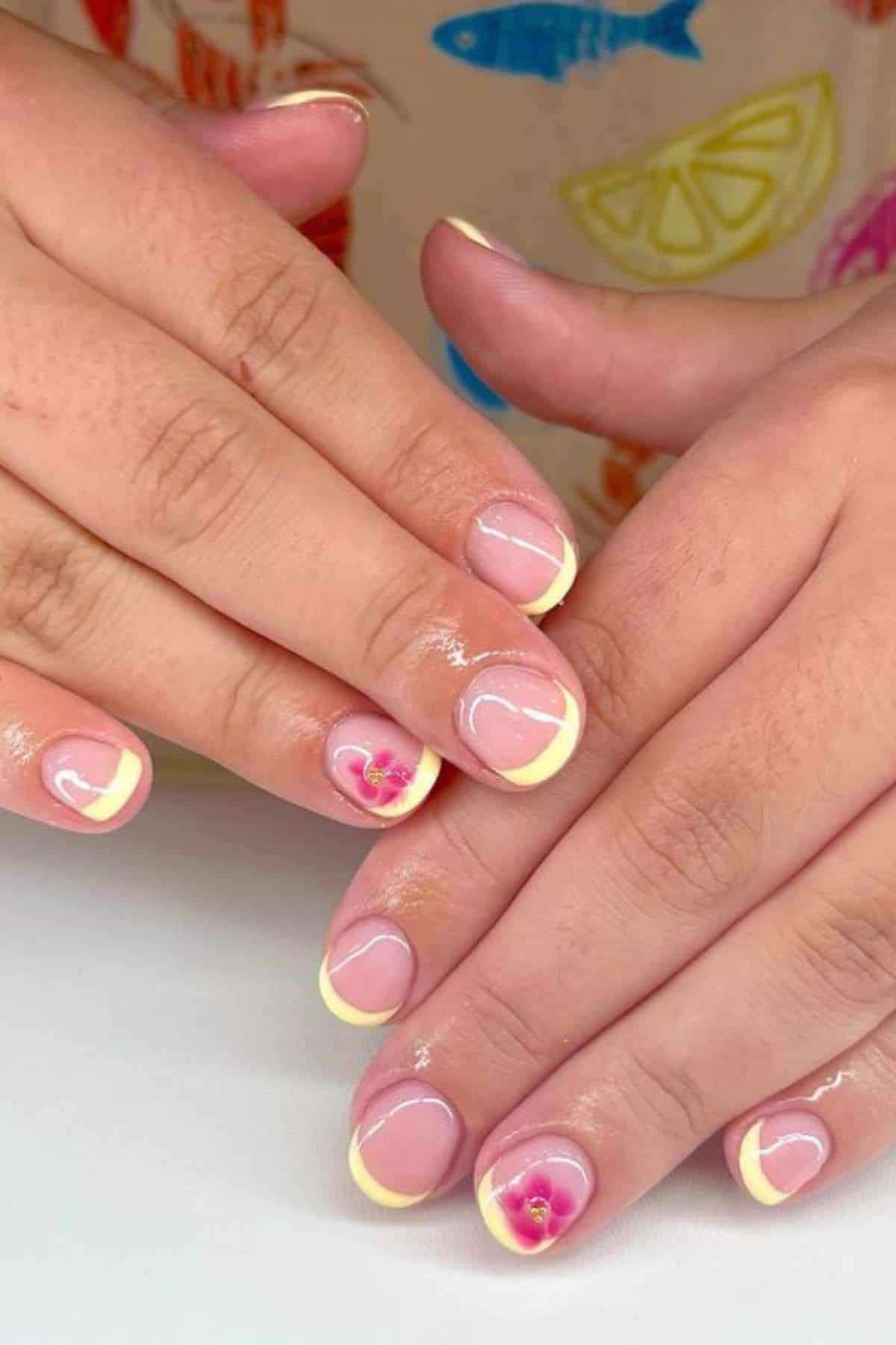 Hands with short nails featuring light yellow French tips and a pink flower accent design on the ring fingers, shown against a white surface.