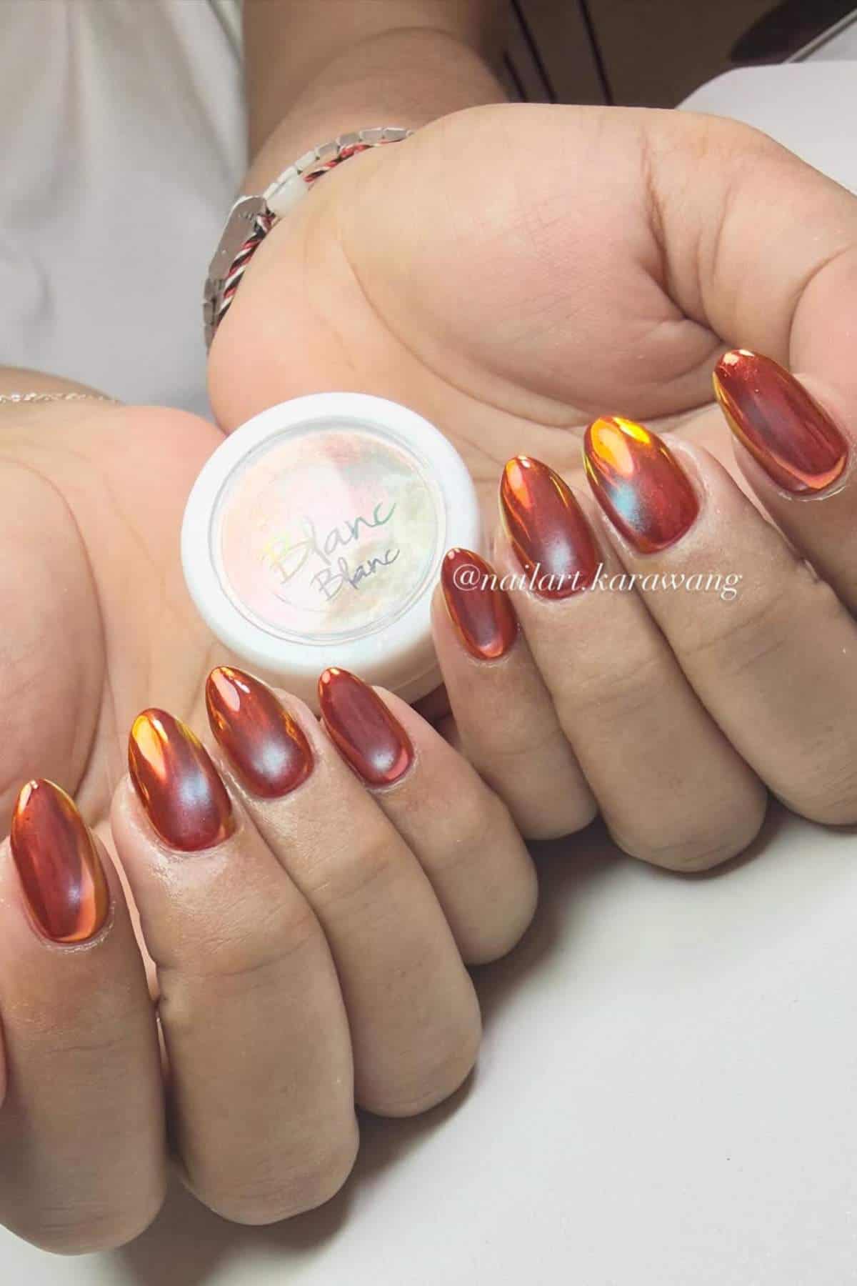 Hands displaying long, almond-shaped nails with a shiny, reflective red chrome finish, holding a small container labeled "Blanc Blanc.