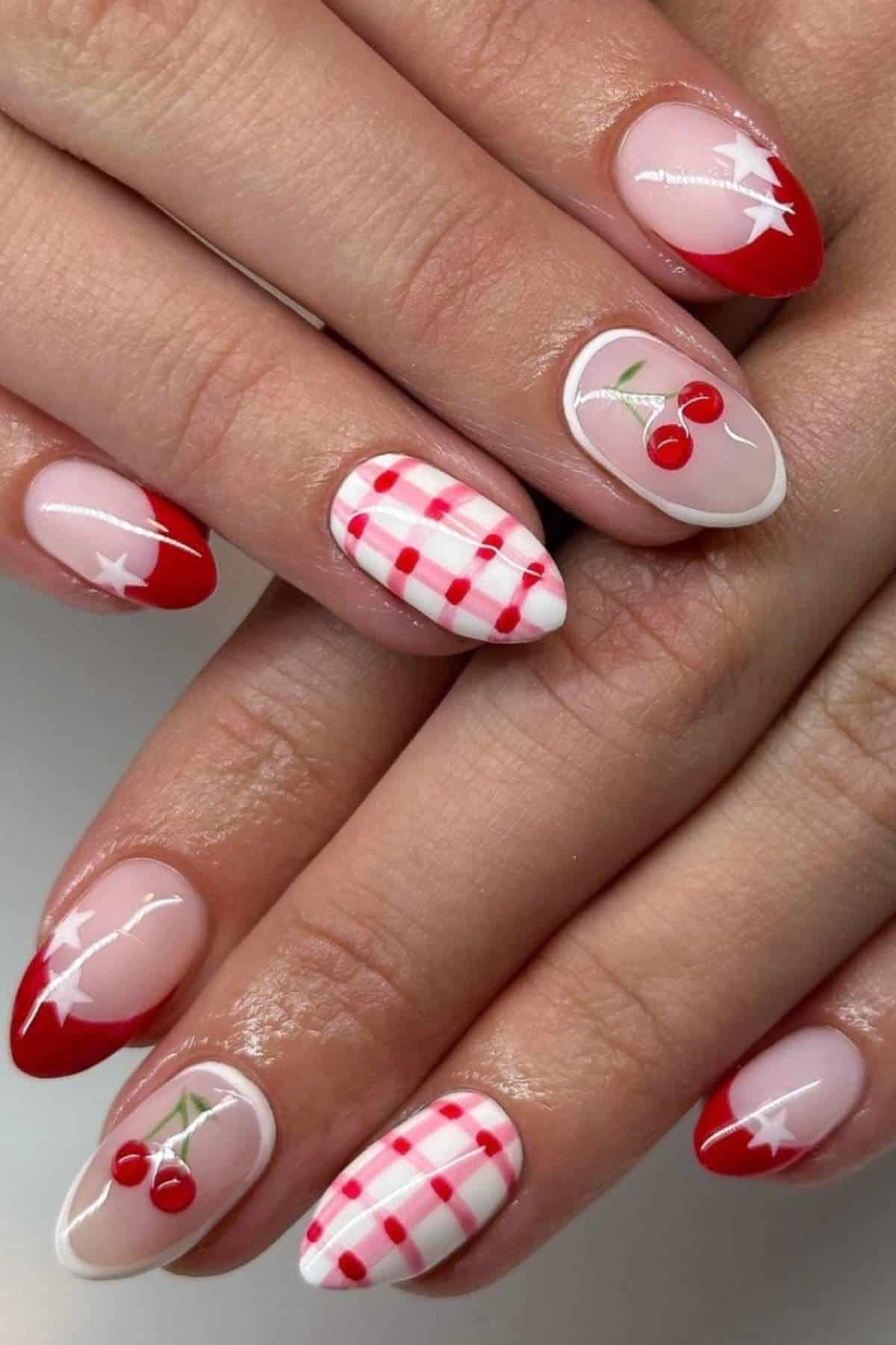 A hand with almond-shaped nails featuring red and white designs, including cherries, red tips, stars, and a red gingham pattern.