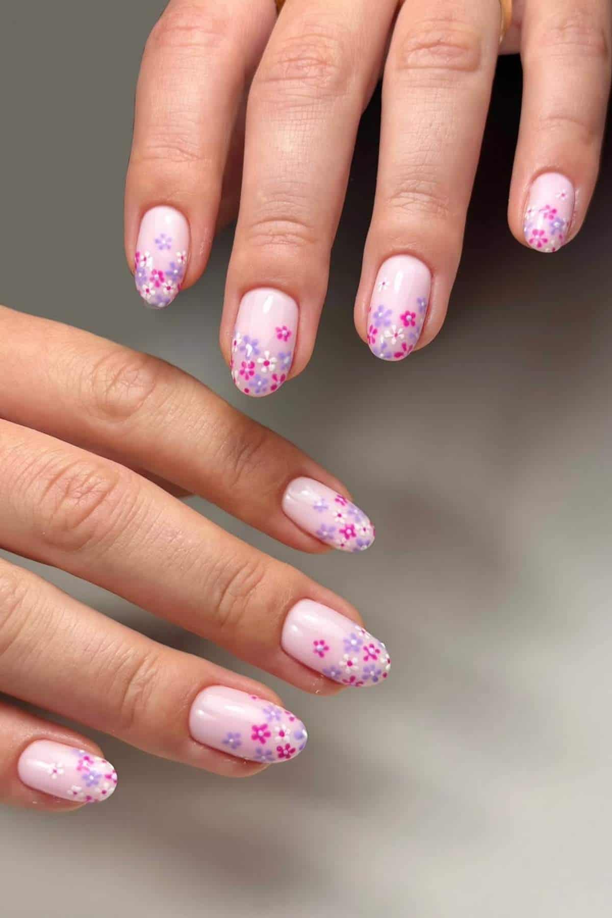 A hand with light pink manicure featuring small purple and pink flower designs on the tips of oval-shaped nails.