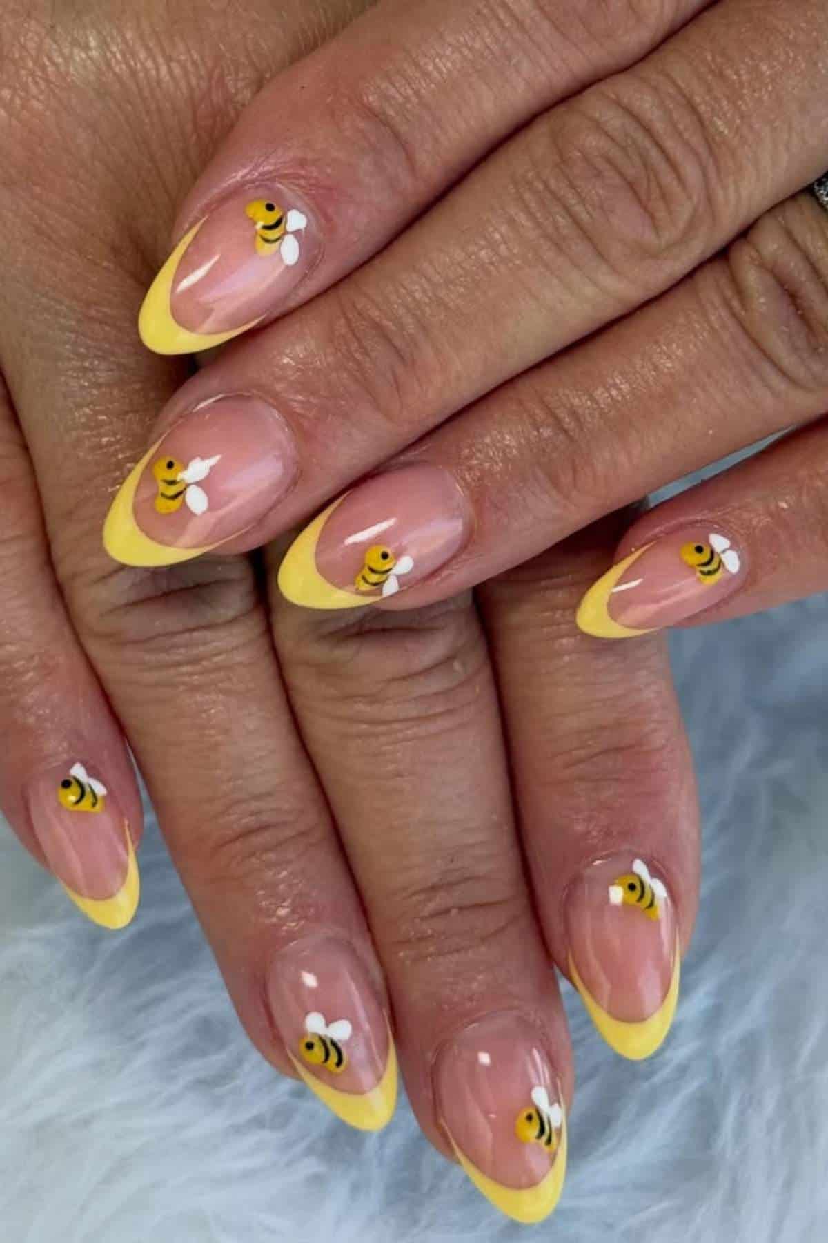 Hands with almond-shaped nails featuring yellow French tips and painted cartoon bees with white wings on a clear base.