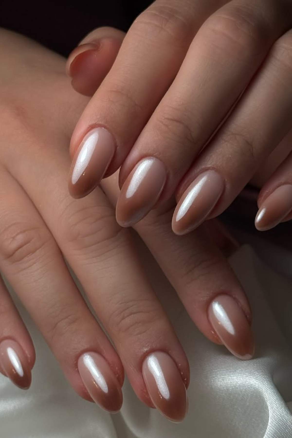 Hands with almond-shaped nails painted in a glossy, pearl nude shade are resting on a white satin fabric.