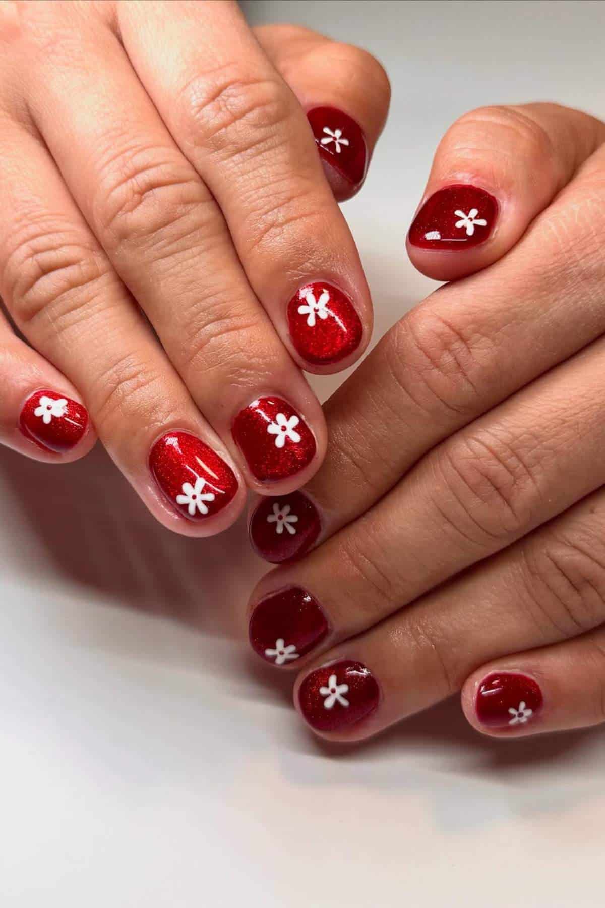 Hands with short, rounded nails painted red and decorated with white flower designs on each nail.