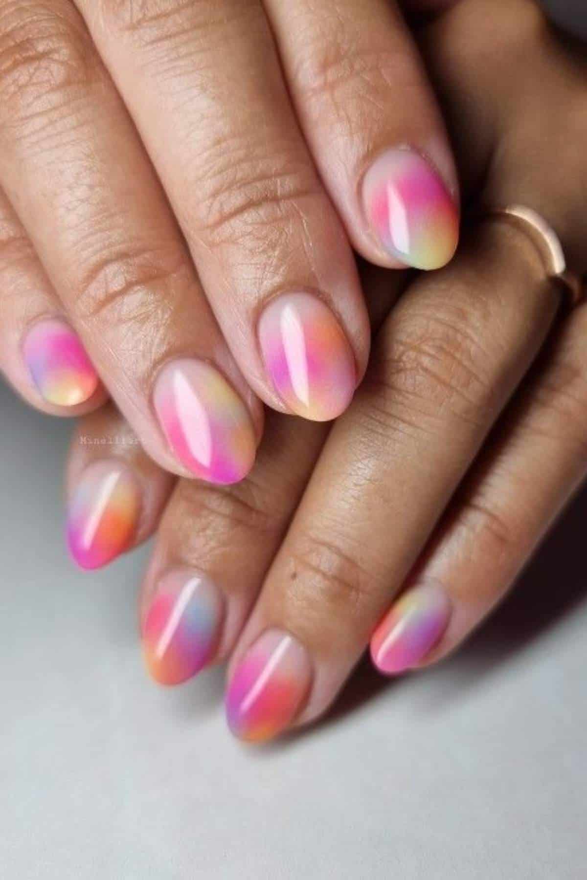 Close-up of hands with short, almond-shaped nails featuring a smooth pastel gradient in pink, purple, orange, yellow, and blue shades, and a simple gold ring on one finger.