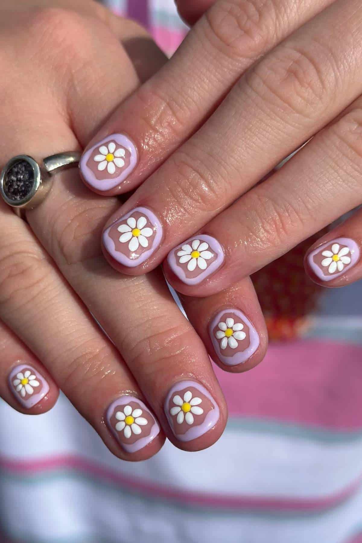 Hands with short nails painted light pink, each nail featuring a white and yellow daisy flower design. A silver ring with a dark stone is worn on one finger.