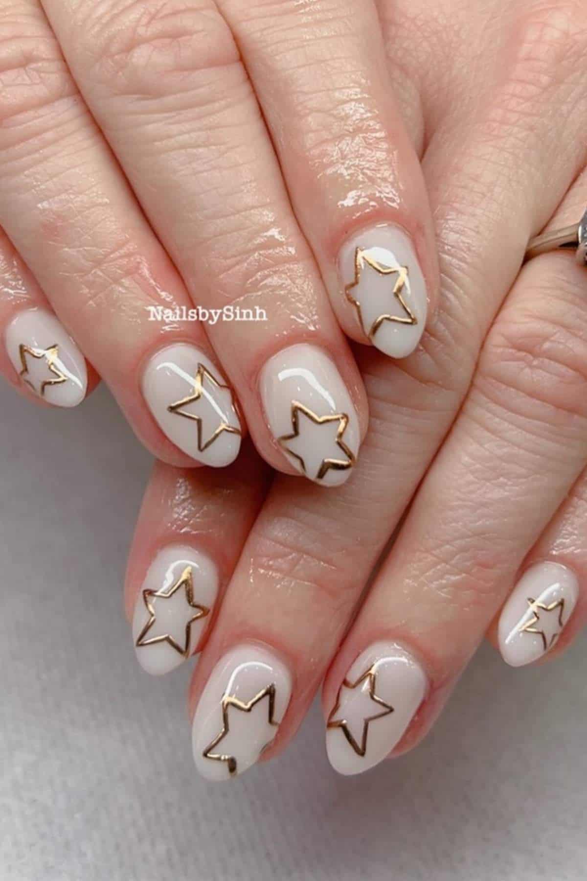 Hands with short, oval-shaped nails painted white and decorated with gold-outlined star designs on each nail.