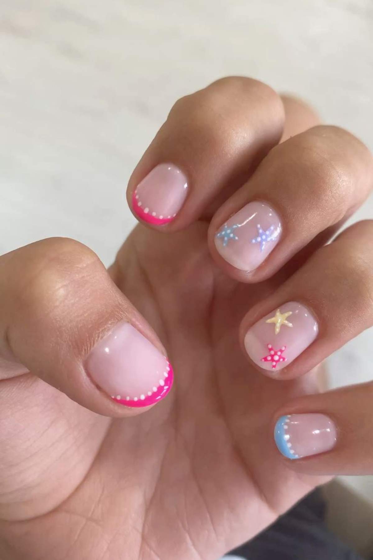 A hand with short nails featuring pink and blue French tips, dotted edges, and small star and dot nail art designs on a light, natural base.