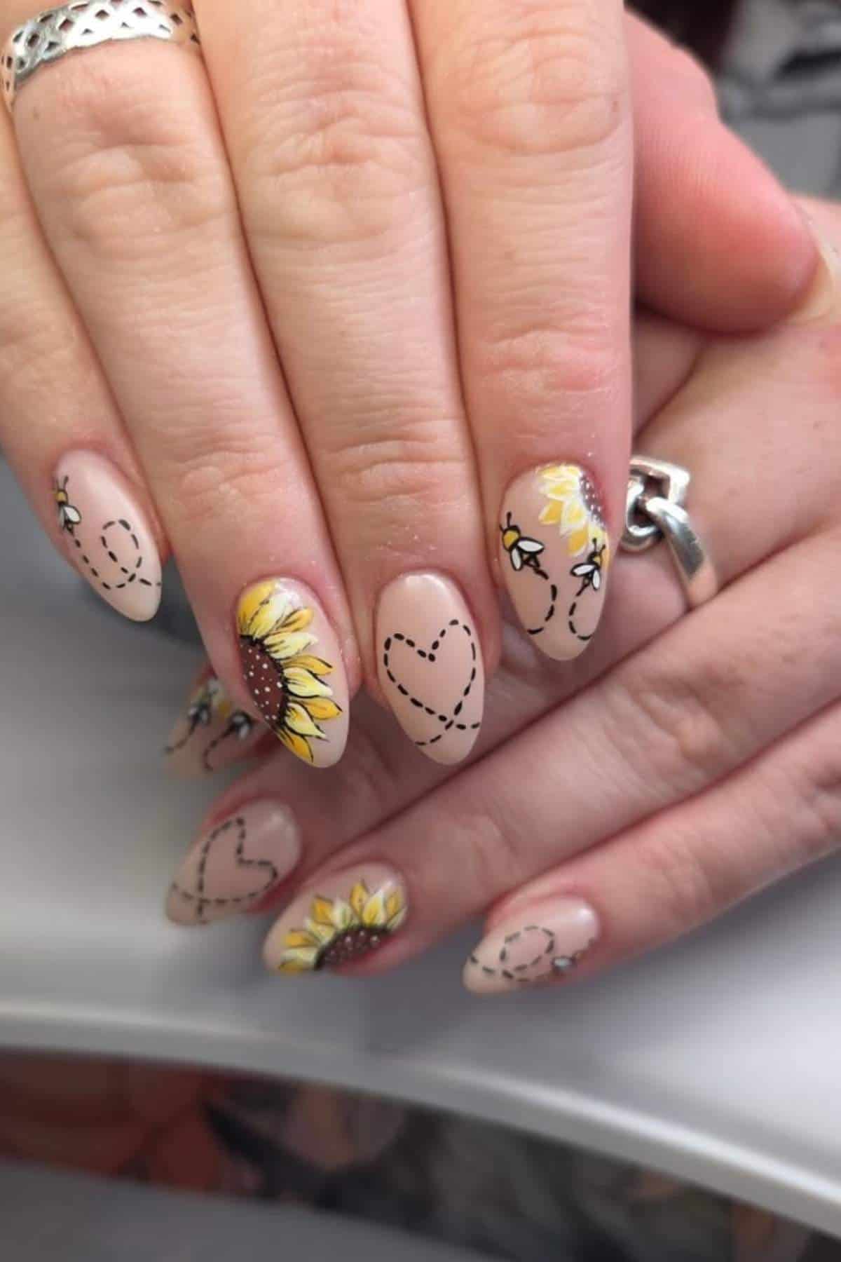 Hands with long almond-shaped nails, featuring sunflower designs and black dotted-line hearts on a nude base.