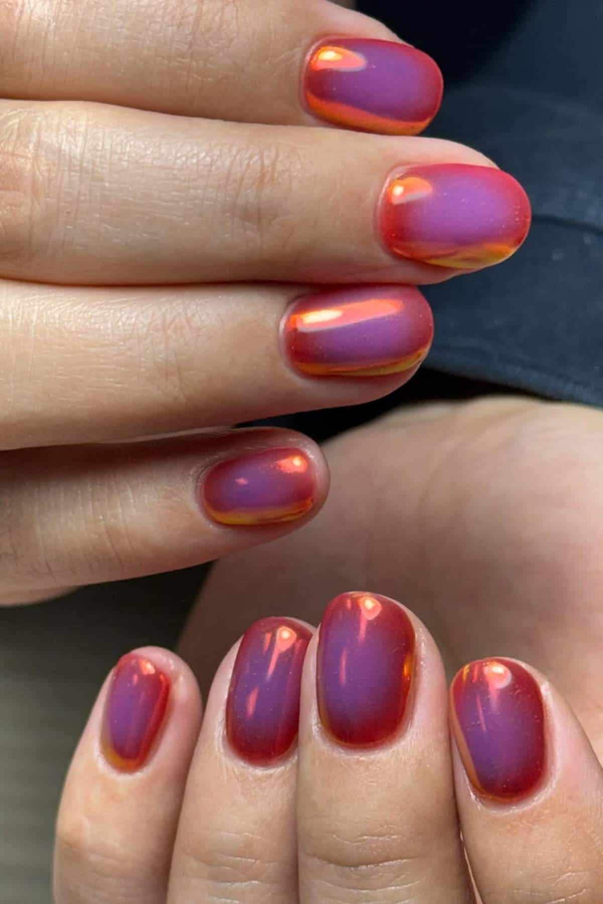 Close-up of hands with short, oval-shaped nails painted in a glossy, iridescent red and purple gradient.