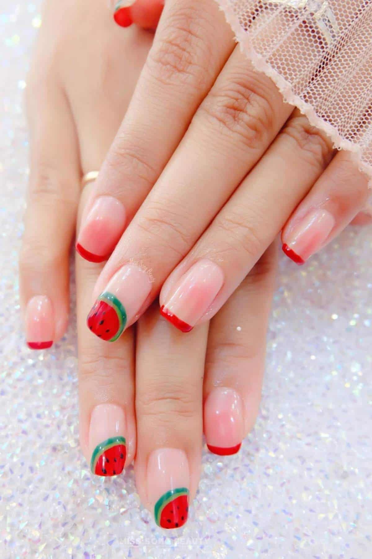 Hands with manicured nails featuring a watermelon design on two nails and red tips on the others, displayed on a sparkling surface.
