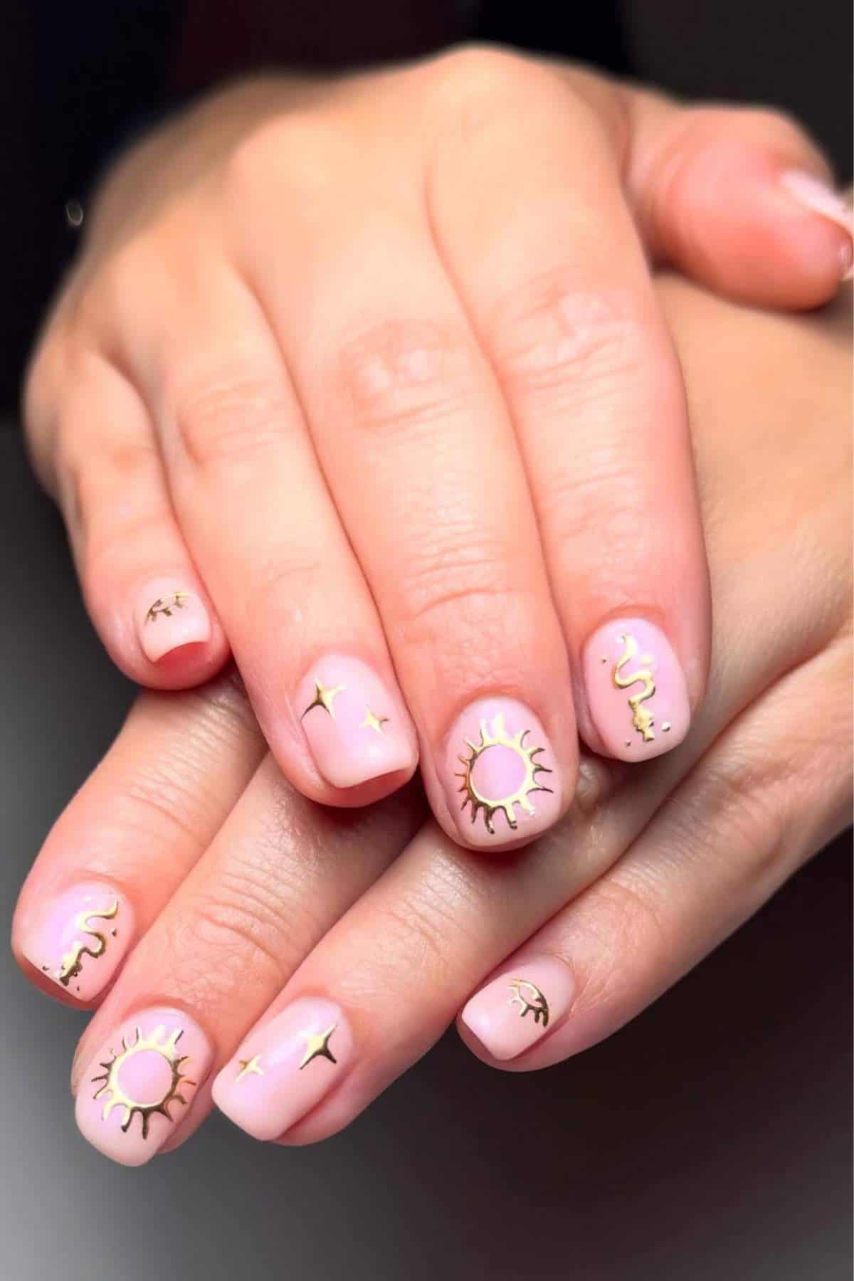 Hands with short, rounded nails painted light pink, featuring gold sun, star, and snake designs as nail art.