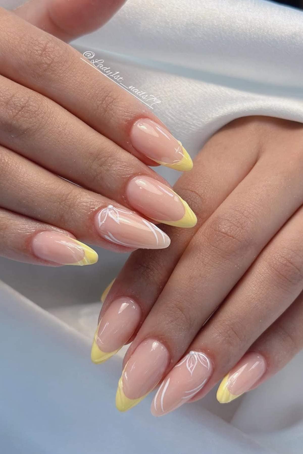 Close-up of hands with almond-shaped nails featuring a glossy nude base and pastel yellow French tips, resting on a white fabric background.