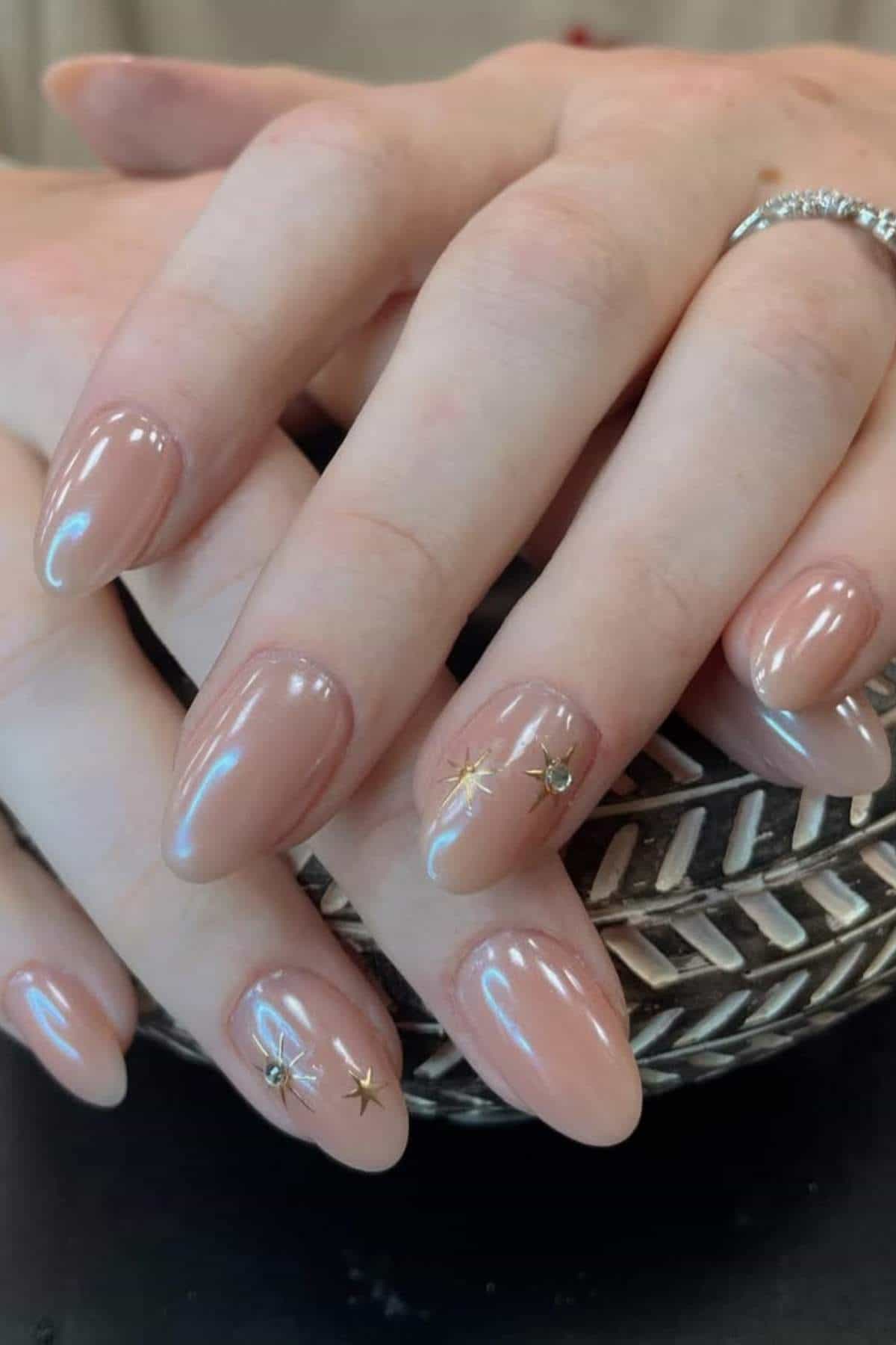 Hands with almond-shaped nude nails, featuring small gold star embellishments on a few nails. One hand wears a silver ring and rests on a patterned object.