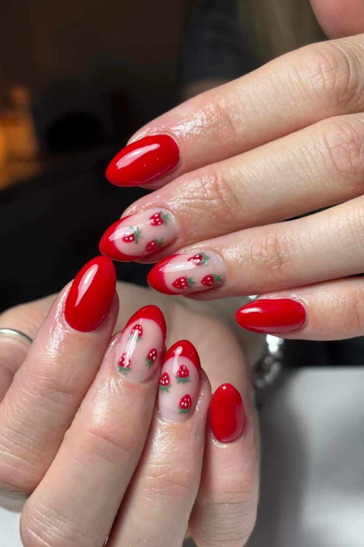 Hands with almond-shaped nails; some nails are painted glossy red, while others have a clear base with small red strawberry designs and green leaves.