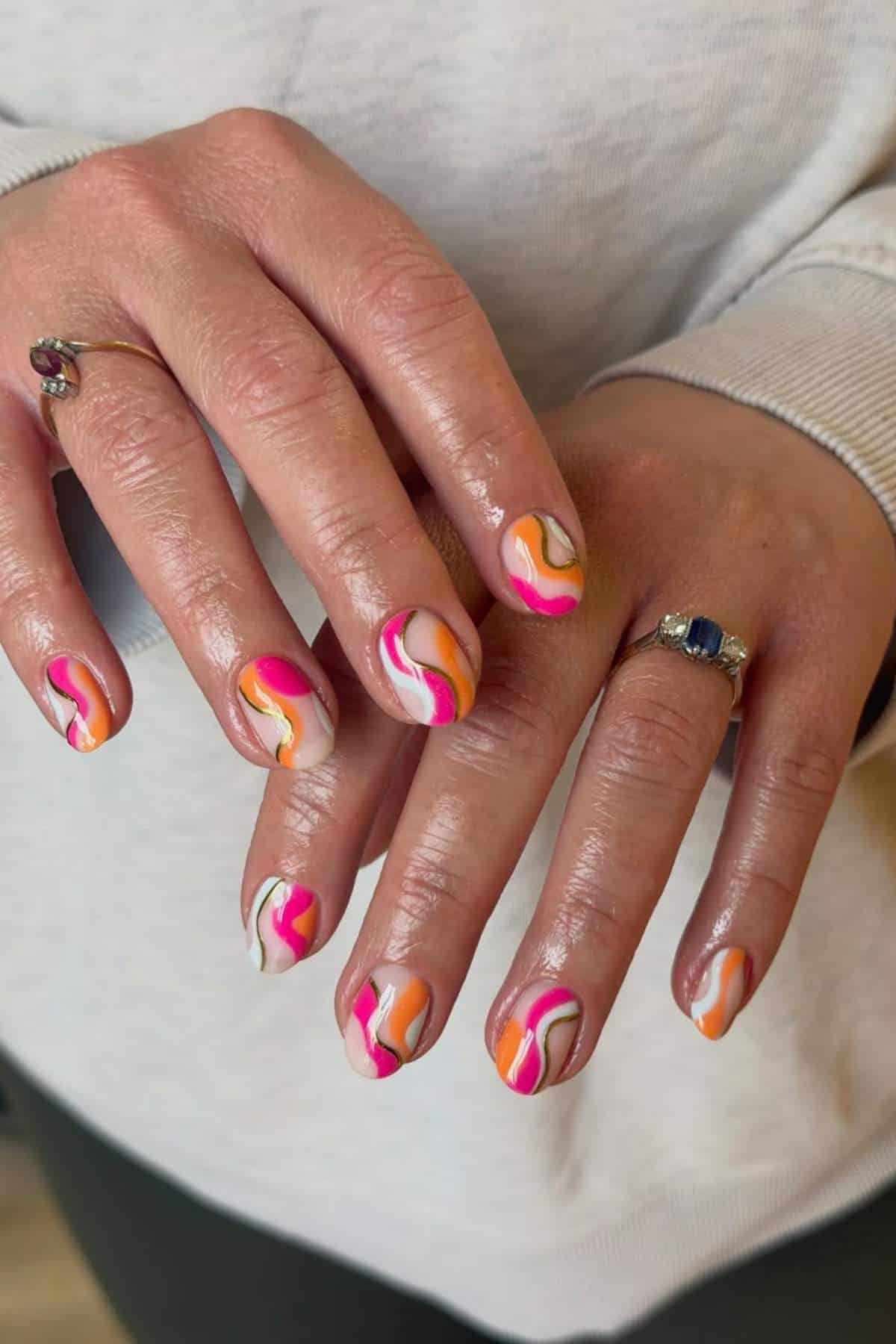 Hands with neatly manicured nails featuring a colorful, wavy abstract design in pink, orange, yellow, and white. The person is wearing two rings and a light-colored long-sleeve top.