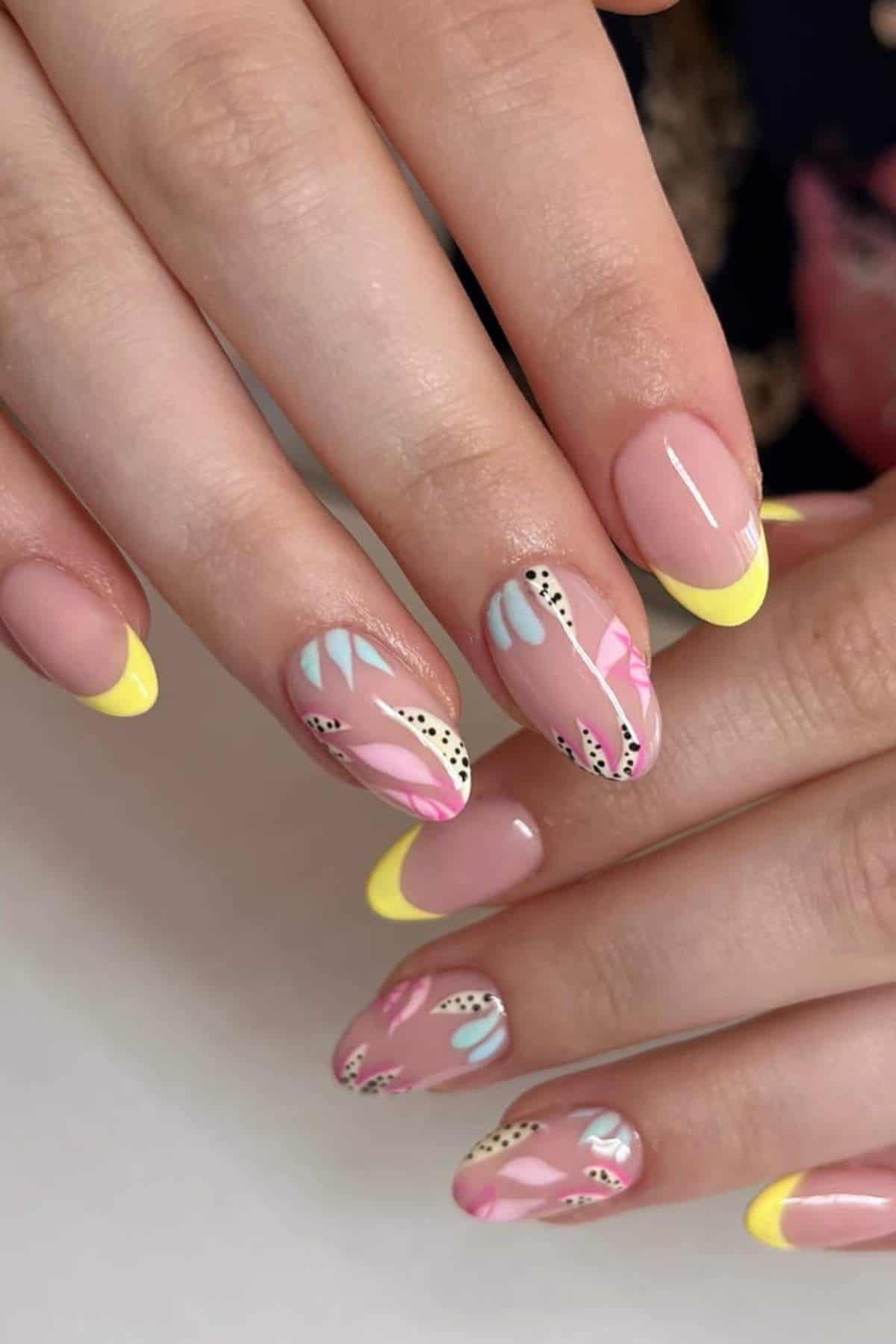Hands with almond-shaped nails featuring yellow French tips and abstract pastel designs with black dots on a nude base.