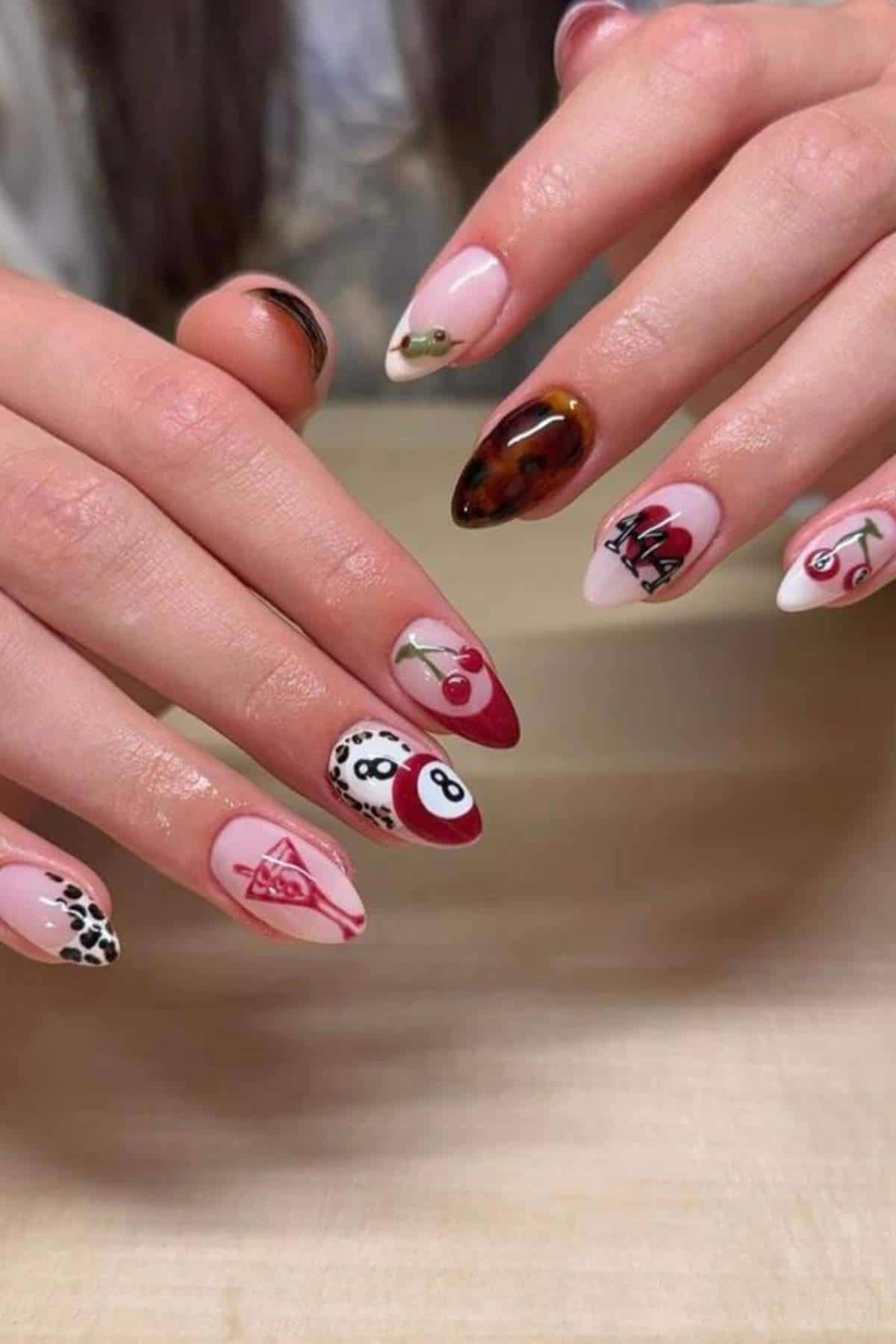 Hands with long, almond-shaped nails featuring various nail art designs, including cherries, the number 8, cow print, flames, and geometric patterns in red, black, and white.