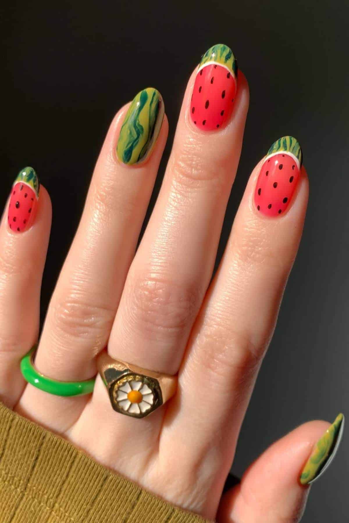 A hand with nails painted to resemble watermelon slices, featuring pink tips with black dots and green bases, shown with two rings on the fingers.