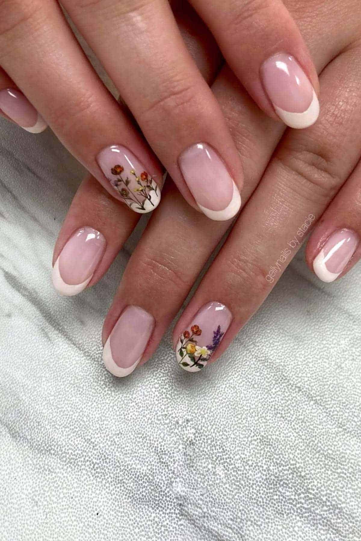 Hands with light pink French tip nails, two nails feature small colorful dried flower designs near the tips.