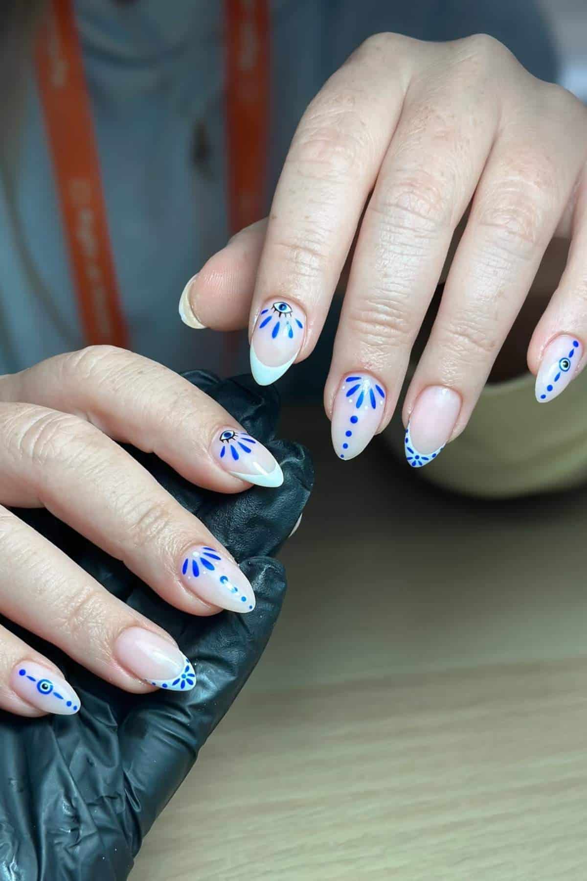 Two hands with almond-shaped nails featuring white polish and blue geometric and floral designs; one hand is held by a person wearing a black glove.