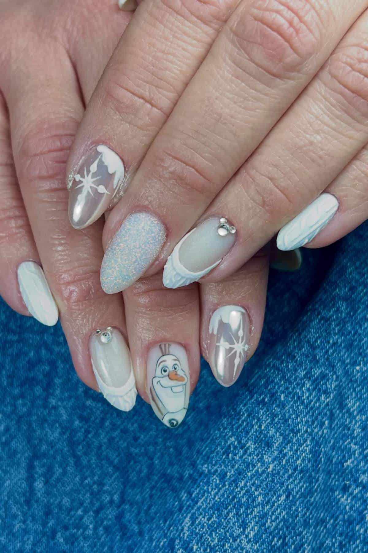 Hands with winter-themed nail art, including snowflakes, rhinestones, glitter, and a cartoon snowman character on one fingernail, resting on blue denim fabric.