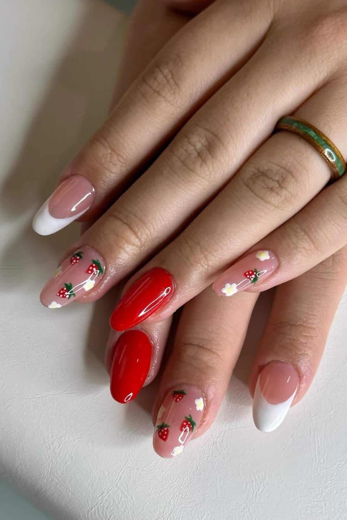 Close-up of hands with manicured nails featuring red polish, French tips, and small strawberry and flower designs on a white surface.