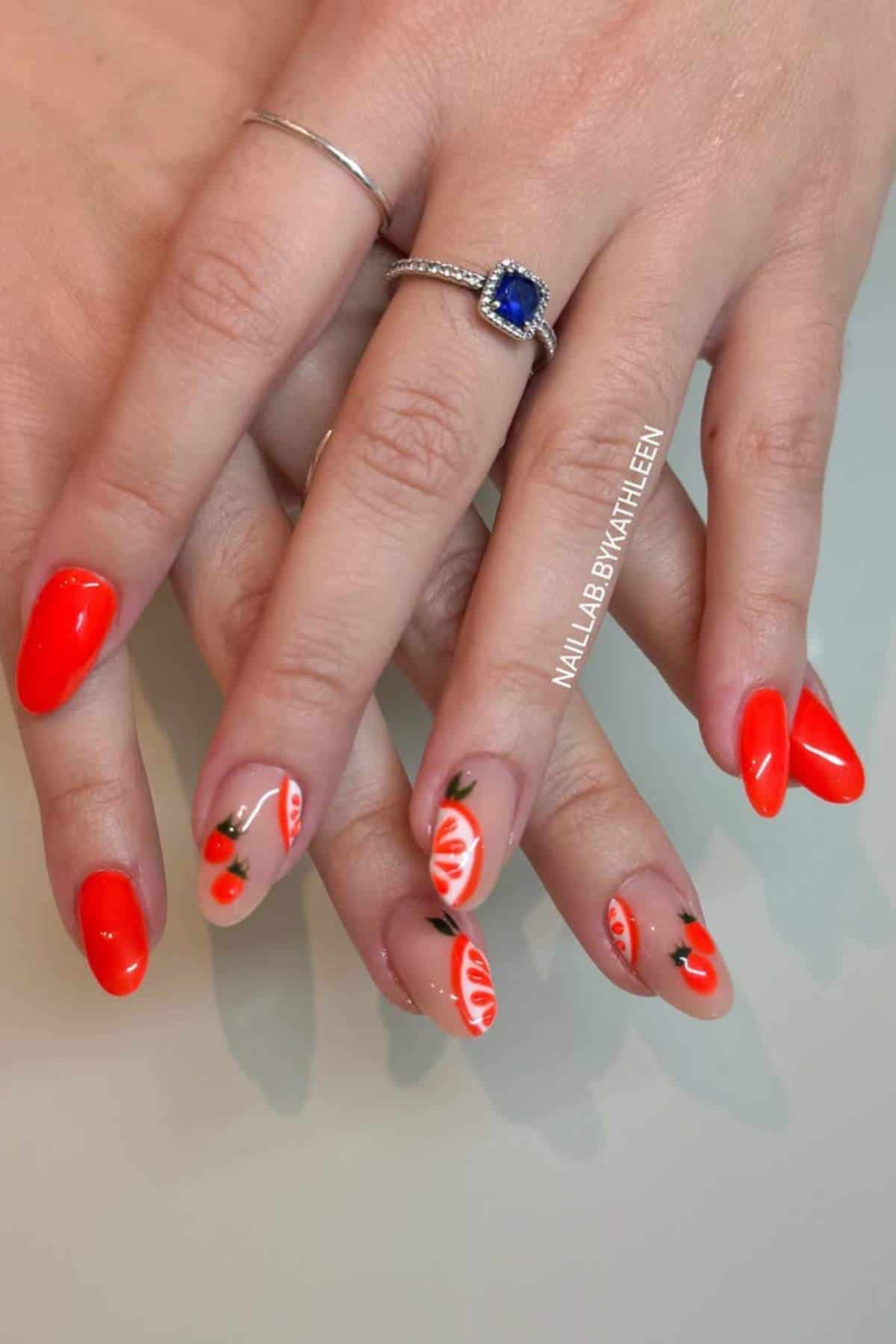 Two hands with oval-shaped nails; most nails painted bright orange, some feature watermelon slice nail art with green rind and black seeds.