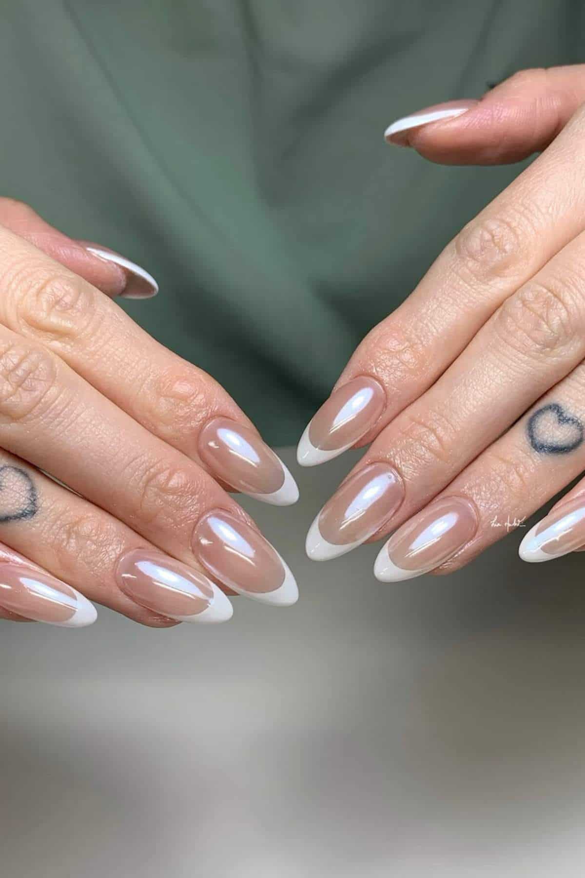 Close-up of hands with almond-shaped nails featuring a pearly nude base and white French tips; small heart tattoos are visible on the fingers.