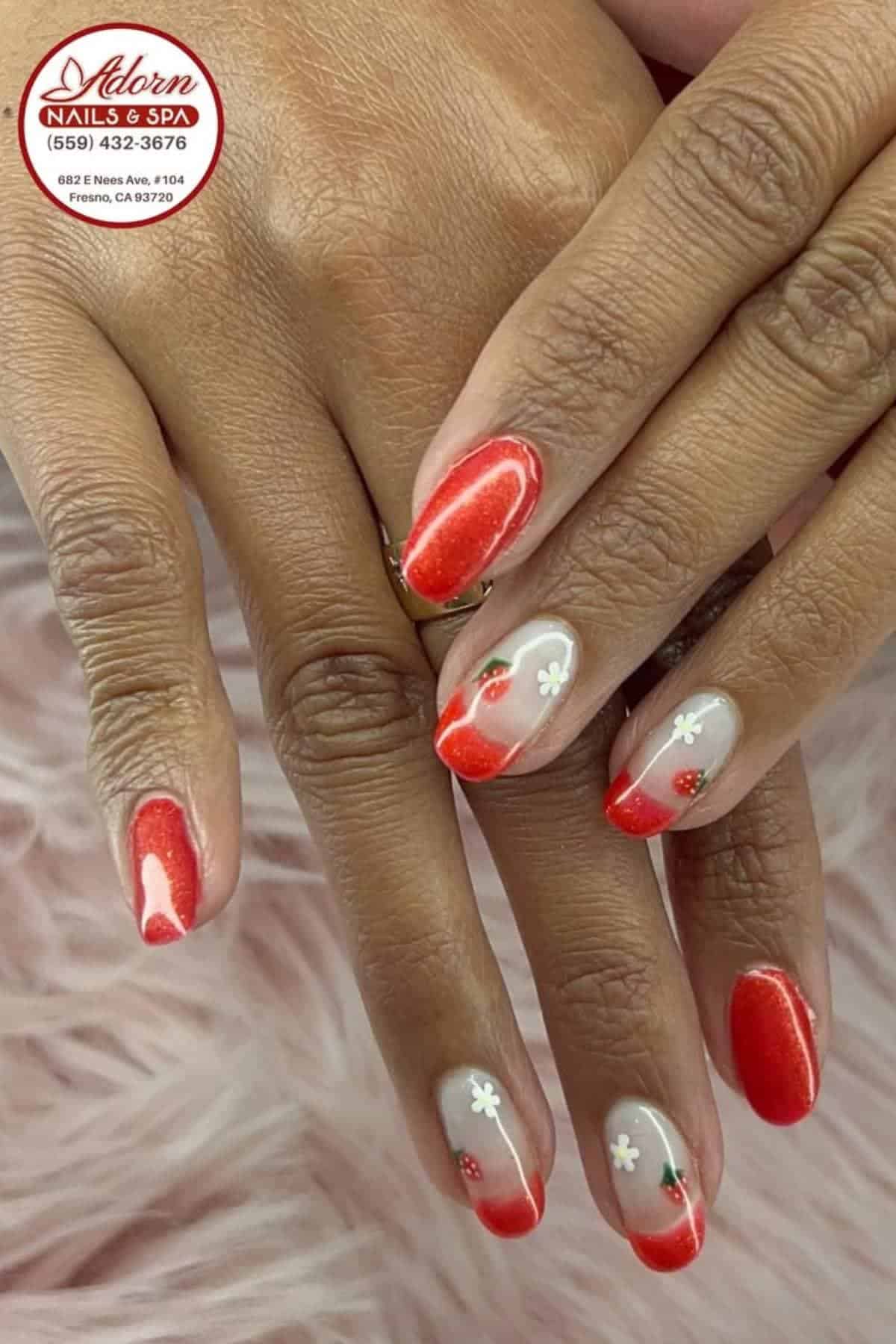 Hands with manicured nails featuring red tips and small white flower designs on some nails, resting on a soft pink surface.