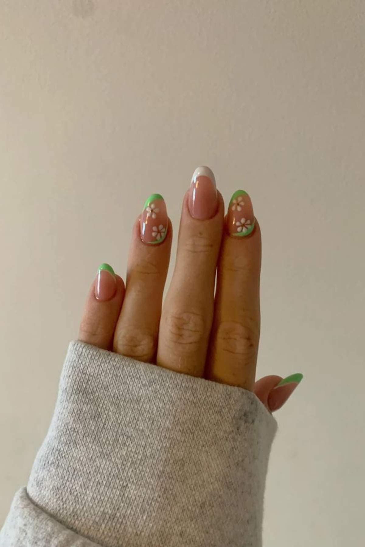 A hand with a light grey sleeve shows nails painted with green French tips and white daisy flower designs on a neutral background.