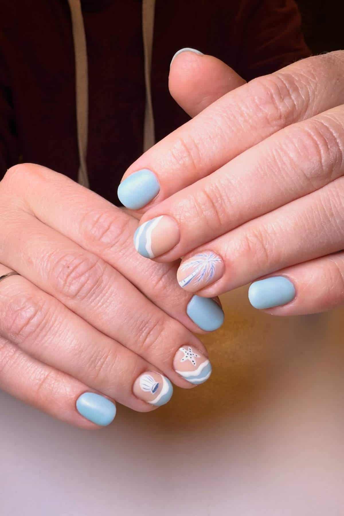 Hands with short, rounded nails painted light blue and decorated with minimalist beach-themed designs, including a shell, starfish, and wave patterns.