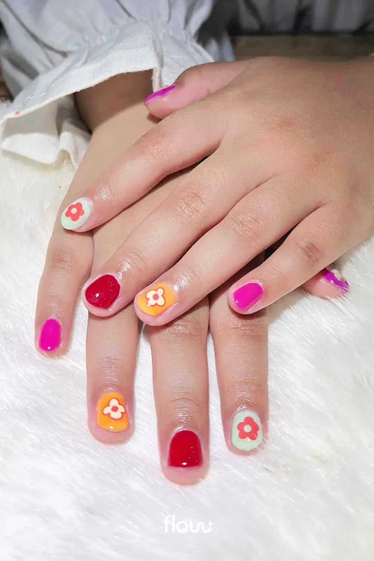 Close-up of hands with manicured nails painted in red, pink, and orange, featuring small floral designs on some nails, resting on a white surface.