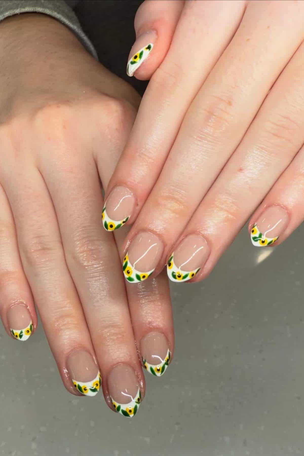 Hands with short, almond-shaped nails featuring a clear base and French tips decorated with white edges and small green and yellow floral designs.