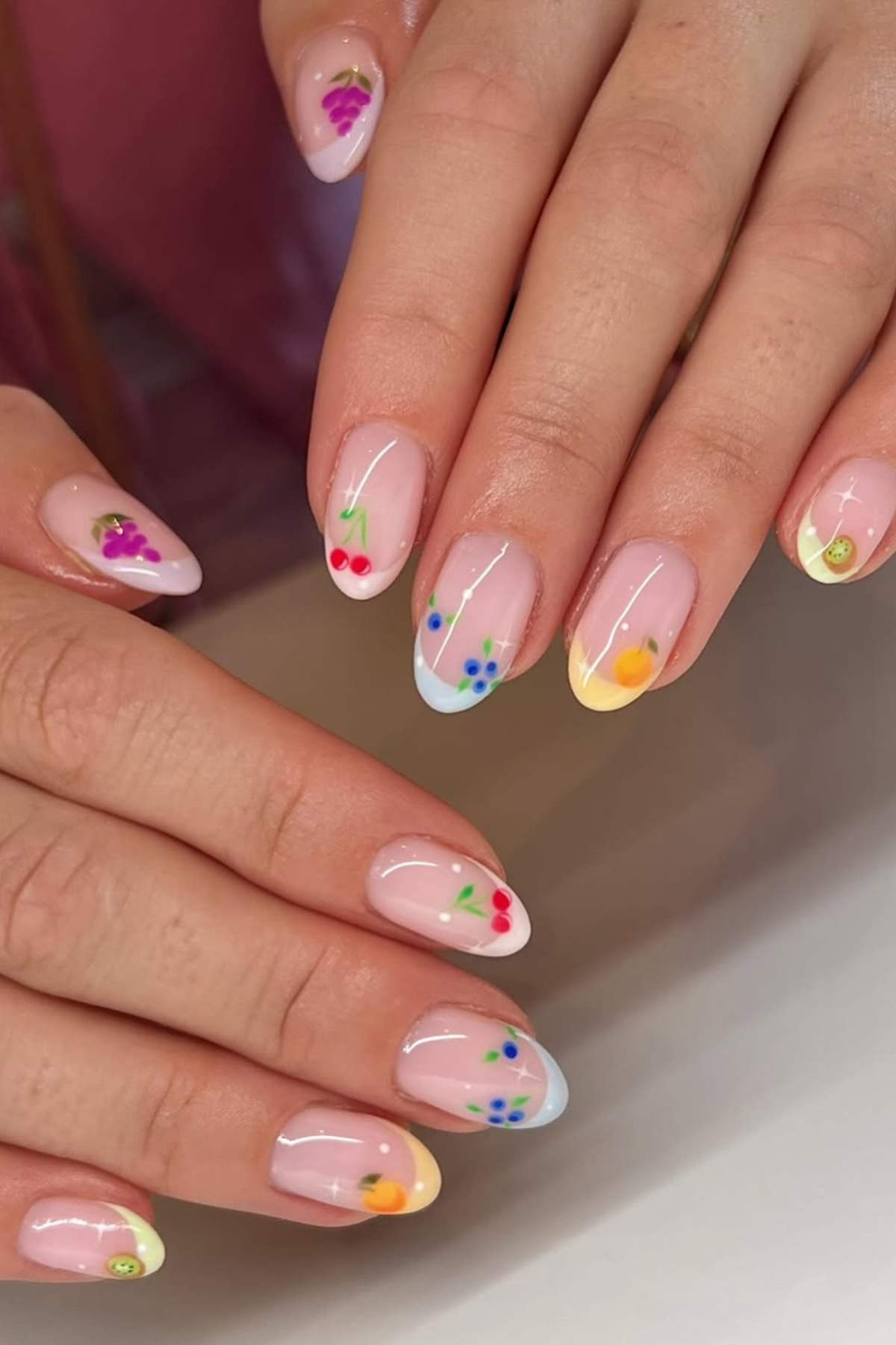 Hands with almond-shaped nails featuring a French manicure style and small, colorful fruit designs painted on each nail.