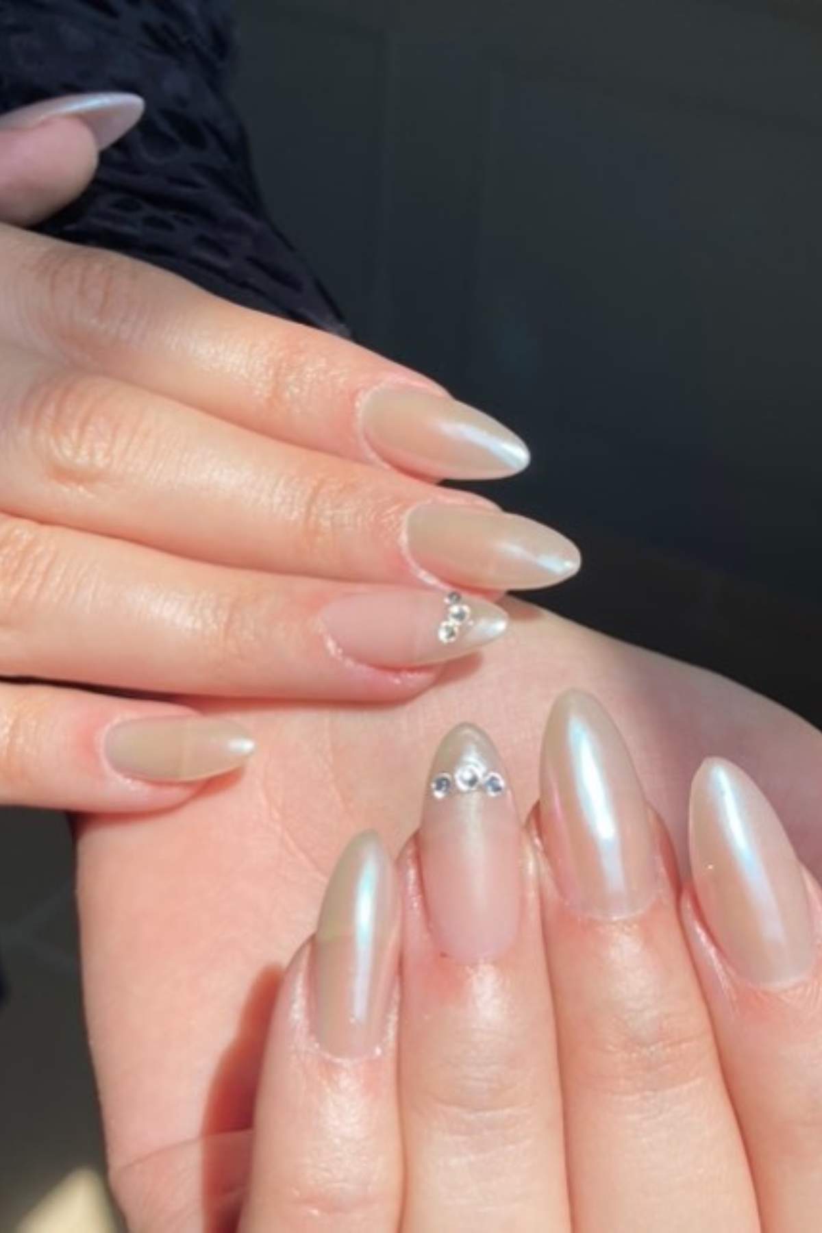 Hands with long, almond-shaped, pearlescent nude nails; one nail on each hand is adorned with small silver rhinestones.