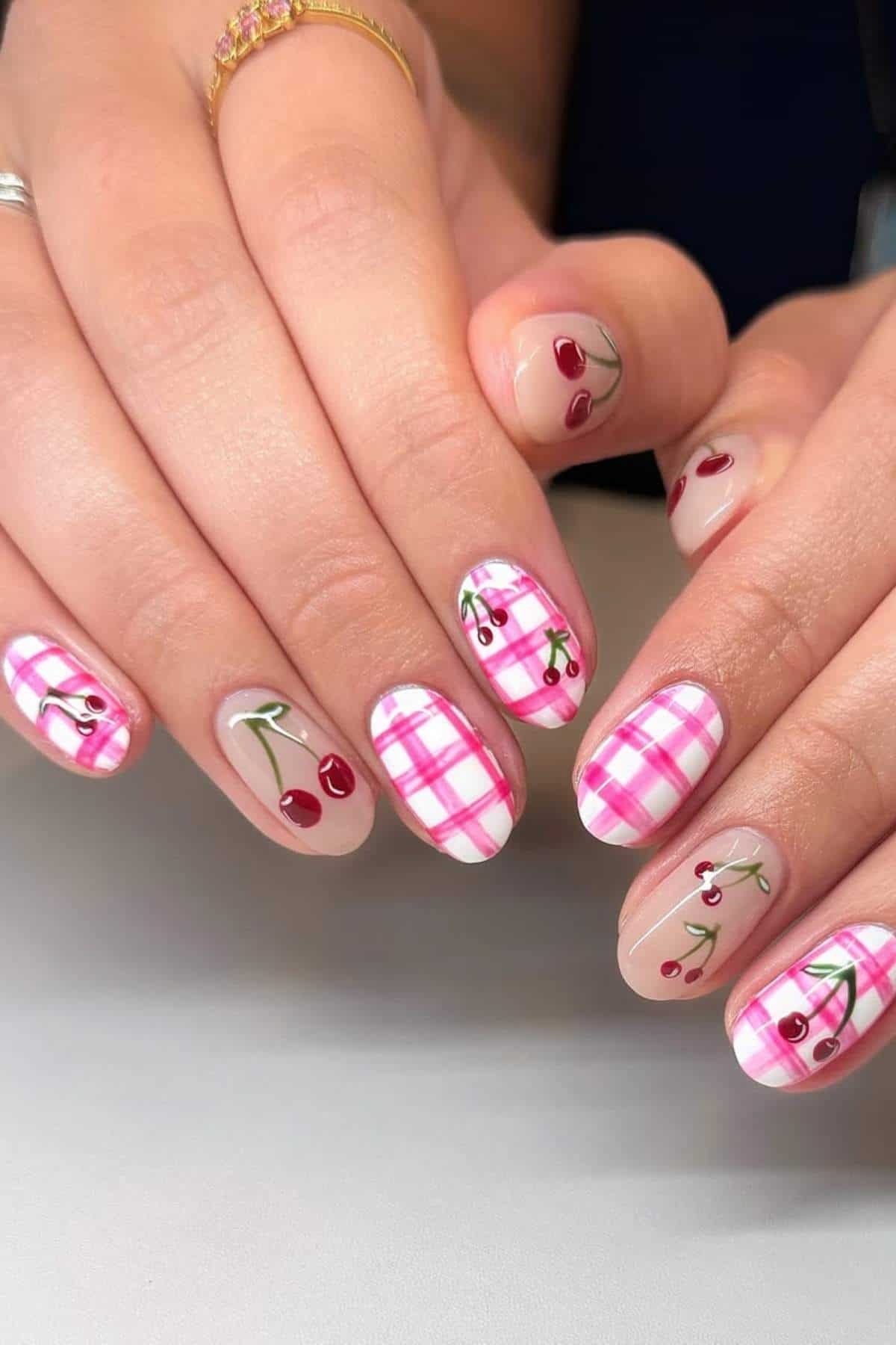 Hands with almond-shaped nails featuring pink plaid and cherry designs on a nude base.