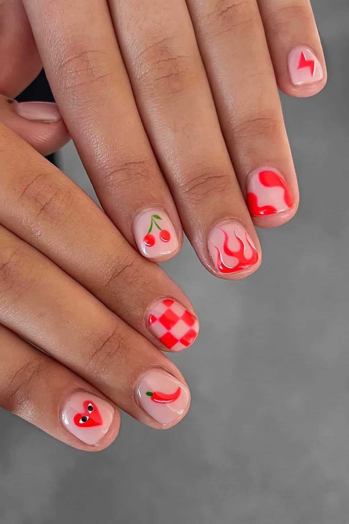 Close-up of hands with short nails featuring various red nail art designs, including cherries, flames, checkerboard, a chili pepper, and abstract shapes on a neutral background.