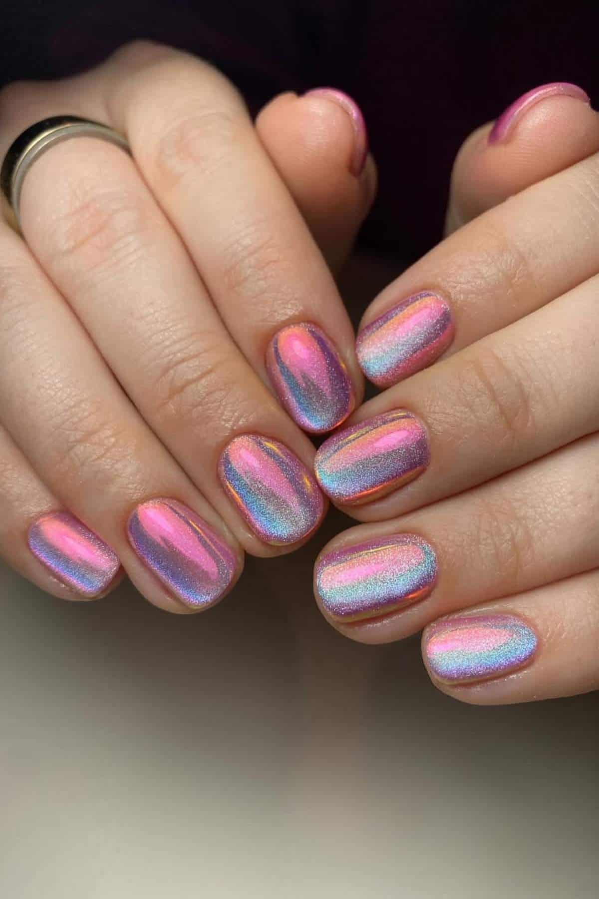 Hands with short, rounded nails painted in a shiny, iridescent pink and blue chrome polish.