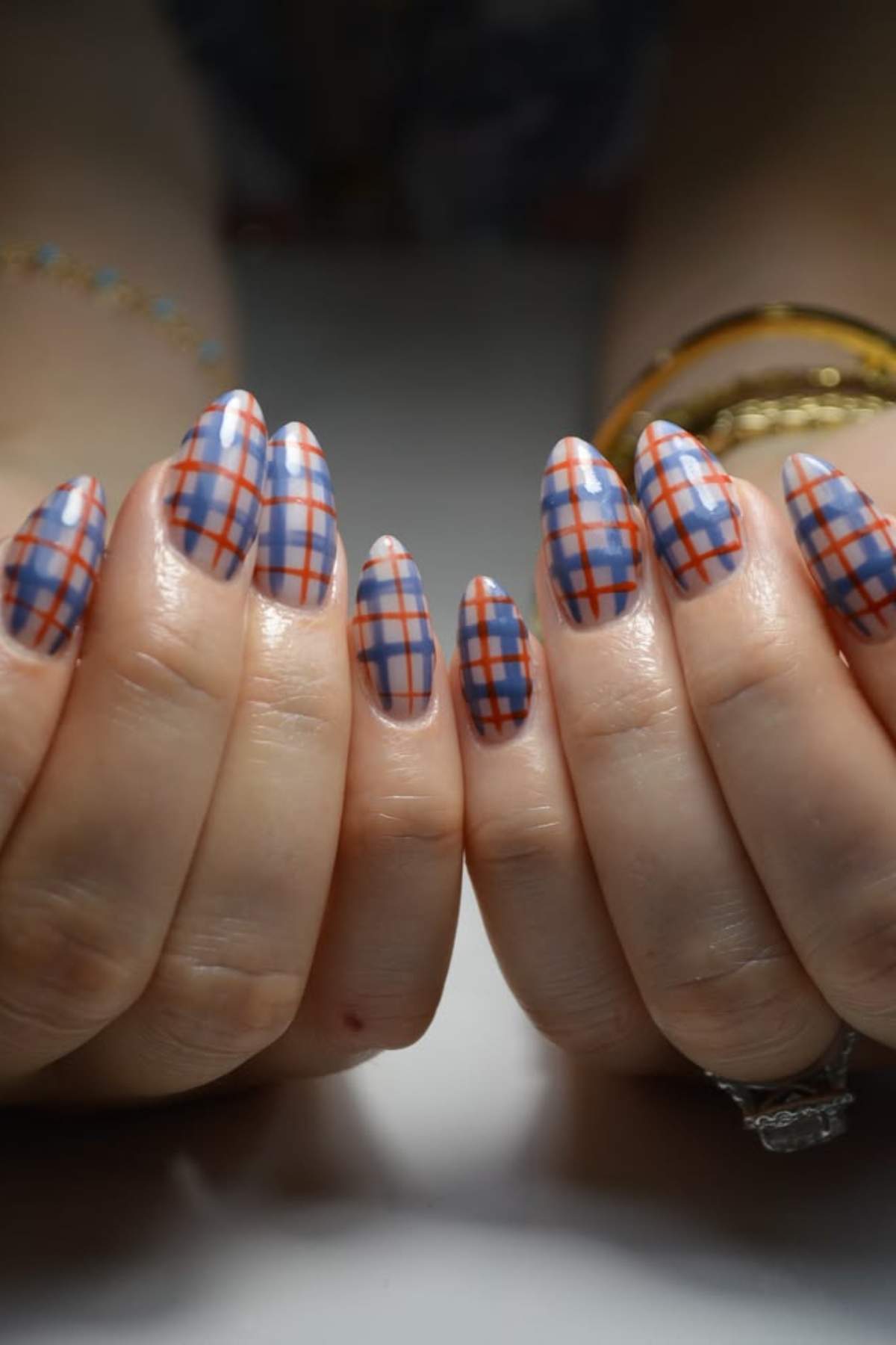 Close-up of hands with long, almond-shaped nails featuring a blue and red plaid design on a nude base.