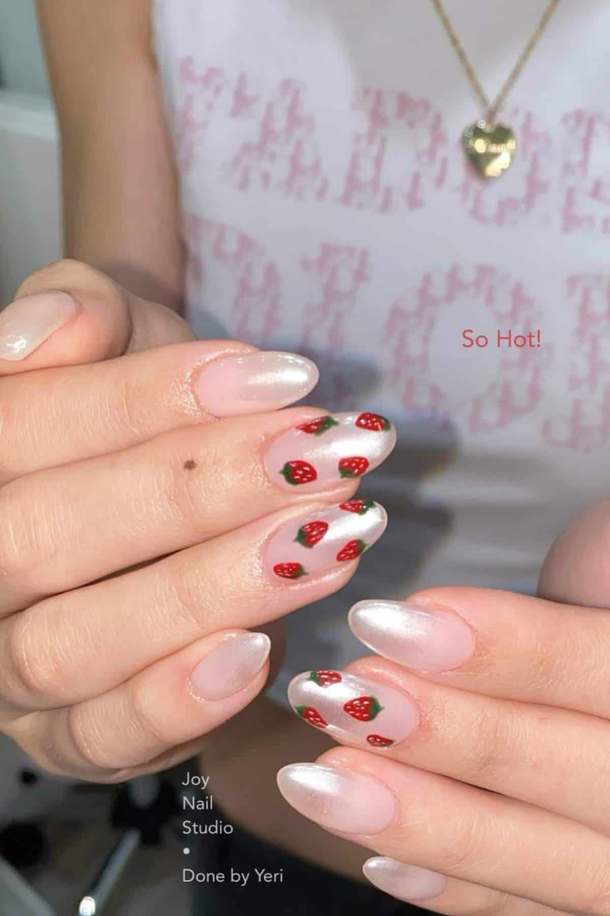 Close-up of hands with almond-shaped nails featuring a shiny, pearlescent finish and some nails decorated with small strawberry designs. Person wears a gold heart necklace and white top.