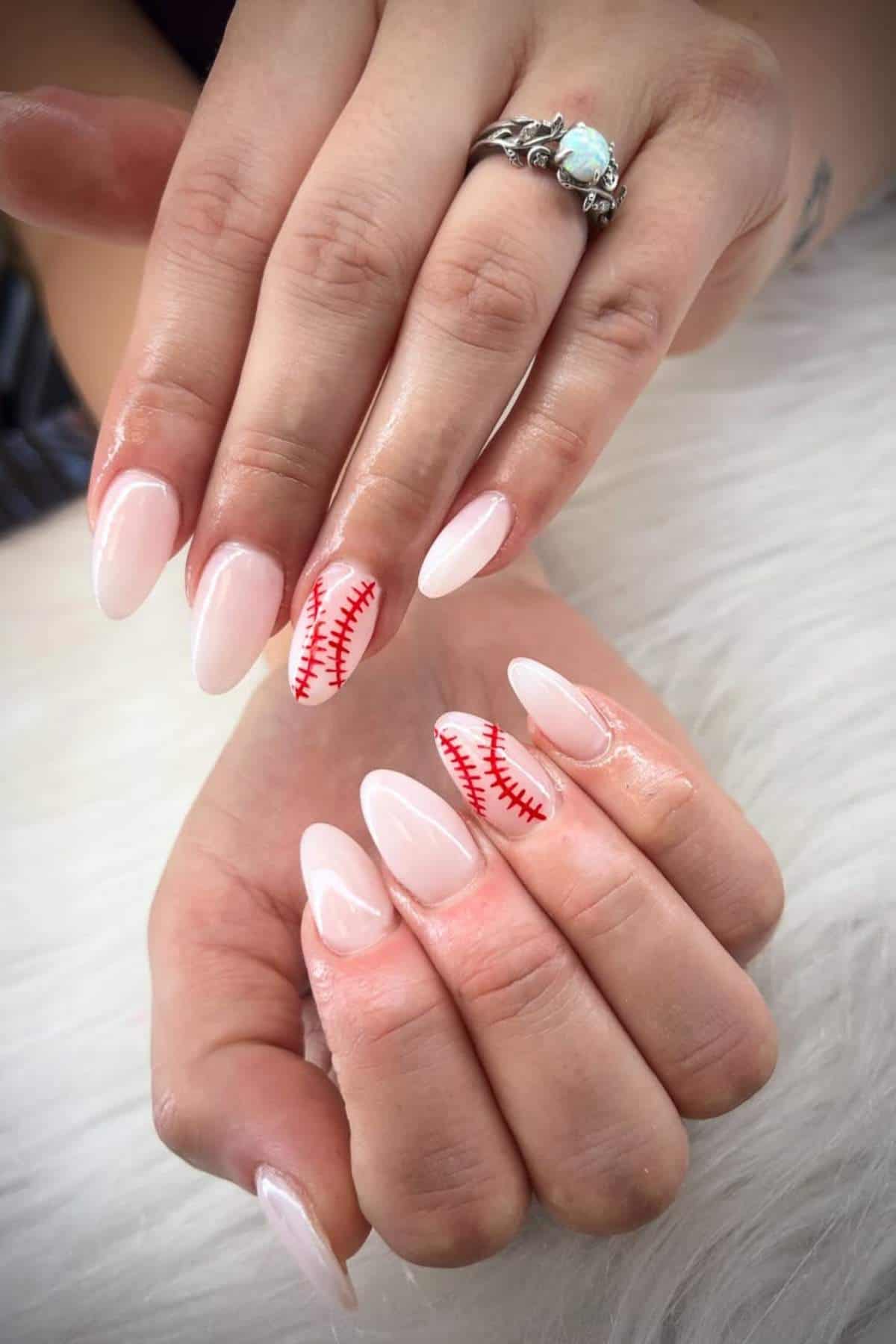 A person with long, almond-shaped, pale pink nails; some nails feature red baseball-style stitching designs.