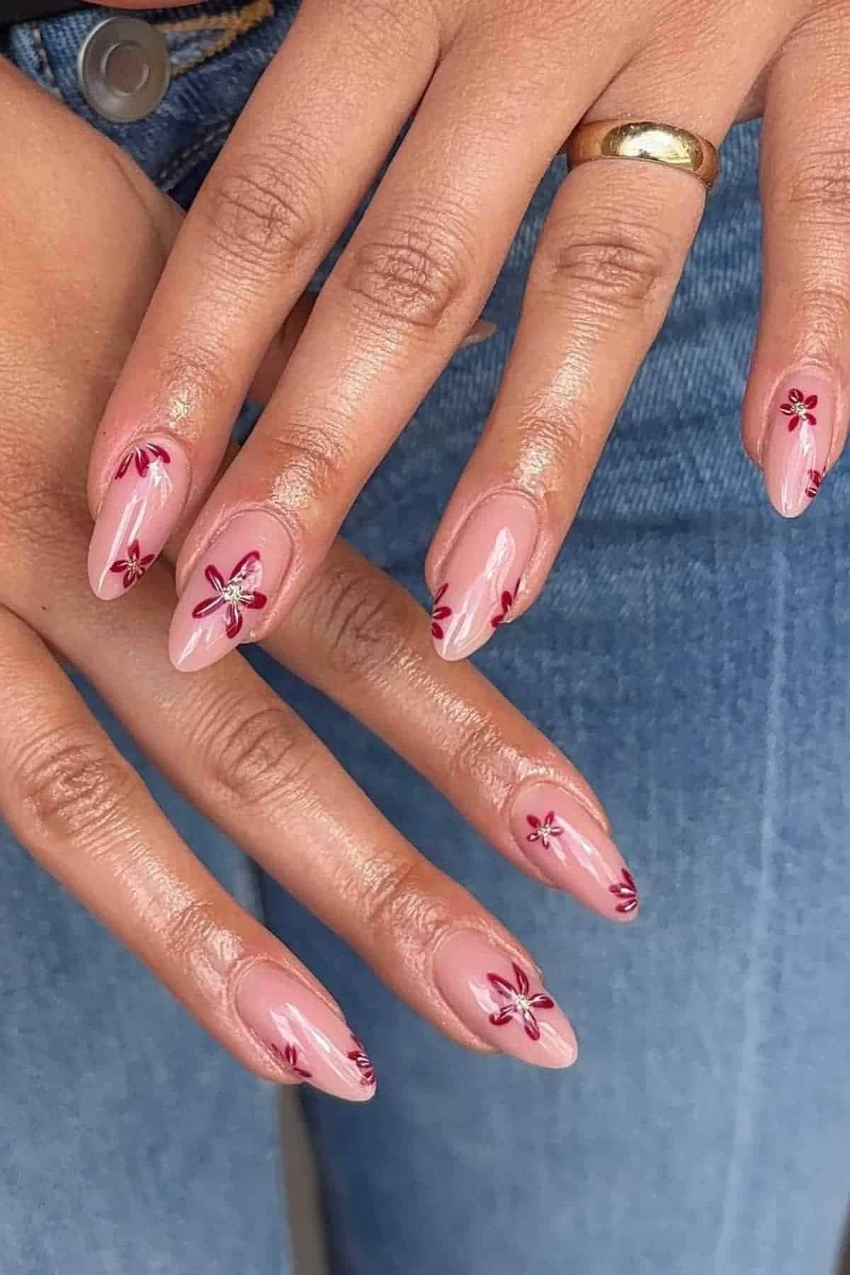 A person with almond-shaped pink nails featuring small red flower designs and rhinestone centers, wearing a gold ring and blue jeans.